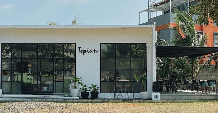 Tampak depan bangunan utama Tepian Café dengan desain minimalis modern berwarna putih, dilengkapi jendela kaca besar dan pot tanaman hias di bagian depan. Area duduk indoor dan outdoor terlihat, dengan beberapa pengunjung yang menikmati suasana di bawah atap kanopi di sisi kanan.
