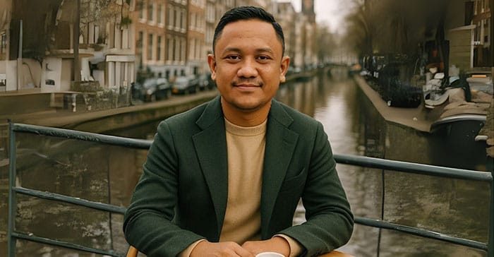 Sigit, Dream Home Specialist dari The Hana Park, duduk santai di sebuah kafe outdoor dengan latar belakang kanal dan bangunan khas Eropa, mengenakan blazer hijau dan sweater cokelat muda sambil tersenyum ke arah kamera.
