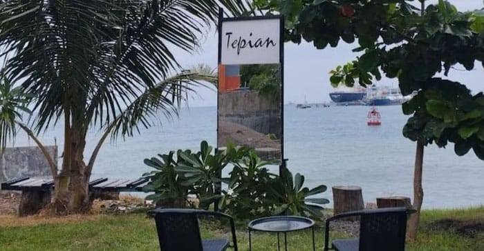Pemandangan area outdoor Tepian Café di tepi laut dengan dua kursi dan satu meja bundar menghadap ke arah laut, dikelilingi tanaman hijau, pohon kelapa, dan latar belakang kapal besar yang bersandar di kejauhan. Sebuah papan bertuliskan "Tepian" berdiri di tengah area sebagai penanda lokasi.