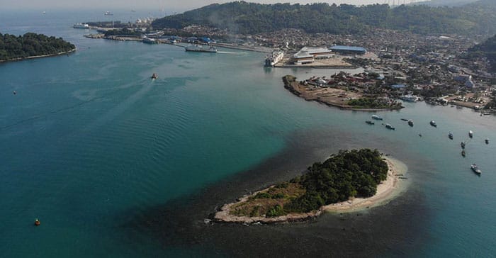 Pemandangan udara kawasan pesisir Cilegon dengan laut biru jernih, pulau kecil berhutan di latar depan, dan area pelabuhan serta kota di kejauhan. Lokasi Tepian Café berada di tepi laut dengan panorama alam yang memukau.